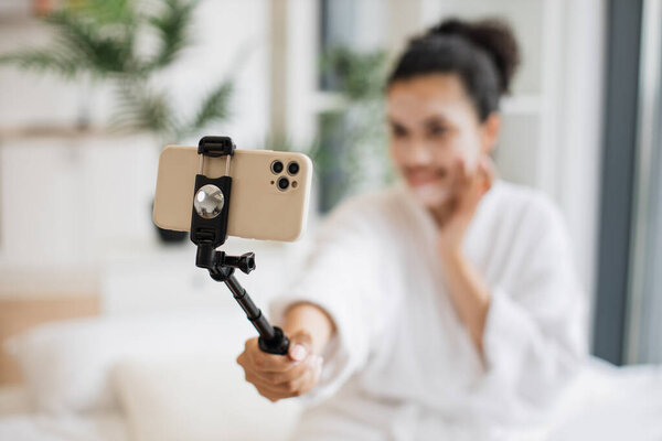Сосредоточьтесь на телефоне расслабленного взрослого блогера в одежде после душа, создавая медиа-контент при нанесении маски, используя косметическое зеркало из лотка стола, сидя на кровати в студии.