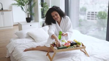 Ellerinde olgun avokado ve kozmetik cam şişeyle gülümseyen bornozlu çekici bayan. Yatakta meyve ve kavanozlarla tepsi masasında dinleniyor. Nazik cilt tedavisi için tedaviyi seçen oldukça yetişkin..