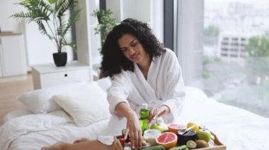 Ellerinde olgun avokado ve kozmetik cam şişeyle gülümseyen bornozlu çekici bayan. Yatakta meyve ve kavanozlarla tepsi masasında dinleniyor. Nazik cilt tedavisi için tedaviyi seçen oldukça yetişkin..