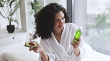 Yatak odasının içinde kozmetik yağ şişesini tutarken elinde avokadoyla kameraya bakan bornozlu güzel sakin bir kadın. Genç bayan yeşil meyvenin büyük cilt faydalarını takdir ediyor..