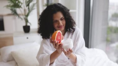 Elinde greyfurt olan bornozlu genç bir kadının ellerine odaklan ve parlak yatak odasının arka planında sağlıklı cilt için ev yapımı krema yap..