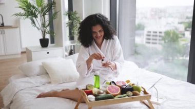 Beyaz bornozlu, gülümseyen bir kadın. Dairenin parlak yatak odasında cilt bakımı için malzemeler hazırlarken manikür yapıyor. Neşeli ev hanımı ojeyle oje sürerek hayat memnuniyetini arttırıyor..