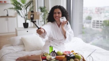 Çok uluslu güzel bir kadın memnun edici, kıvırcık saçlı, sağlıklı ciltli, telefonla poz veren, parlak, modern yatak odası dairesinin arka planında beyaz bornozlu krem süren yaratıcı..