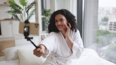 Selfie çubuğunda telefonu olan ve yanağına krem süren çok uluslu genç kadın vlogger 'ı kapatın. Kıvırcık saçlı, taze sağlıklı tenli, sosyal medya için mutlu bir şekilde gülümseyen çekici bir kız..