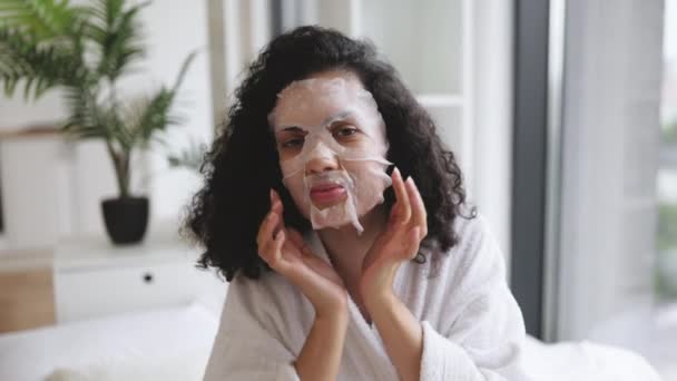 白いバスローブのかなりアフリカの女性は 自宅で自由な時間を過ごしながら 応用紙シートマスクで顔に触れています ベッドルームの内装で美容ケアの手続きに恩恵を受ける若者 — ストック動画