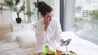 Yatak odasının içinde meyve ve kozmetik ürünlerle yüz maskesi takan bornozlu genç bir kadının yakın çekimi. Yüz kozmetik maskeleri cilt sigortası ile yükleniyor.