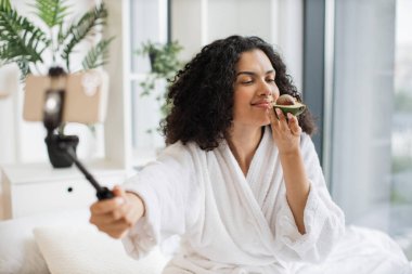 Bornozda çok ırklı kadın sosyal medya etkileyicisi izleyicilere telefonla güzellik ürünleri sunuyor, kokluyor ve avokado, cilt bakımı konseptinin yarısının tadını çıkarıyor..