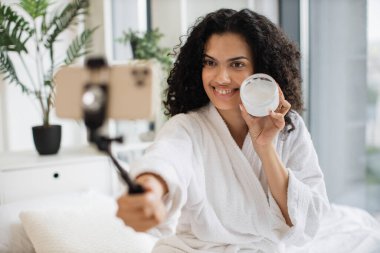 Genç Afrikalı kadın sosyal medya etkileyicisinin portresi bornoz akışı ve izleyicilere telefon ile güzellik ürünleri tanıtmak nemlendirici krem, cilt bakımı konsepti.