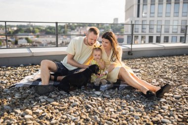 Şehir binasının çatısında el ele tutuşurken battaniyenin üzerinde oturan ve küçük kızına sarılan sevgi dolu ebeveynler. Beyaz kadın ve erkek, bebeğin evcil hayvanlarla ortak bir dil bulmasına yardım ediyor..