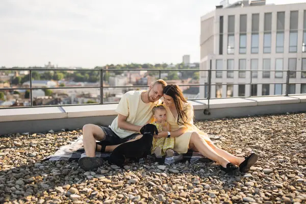 Loving experienced parents sitting on blanket and hugging little daughter while holding hands on roof of city building. Caucasian woman and man helping baby to find common language with pet.