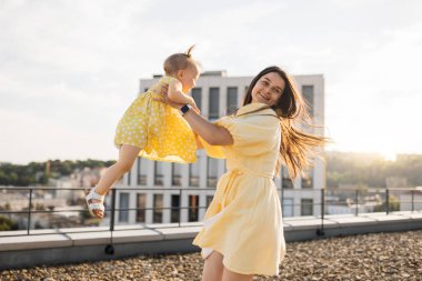 Güzel, gülümseyen esmer, küçük kızını kollarına alıp gökdelenlerin çatısında dönüyor. Yazlık sarı elbiseler giyen neşeli yetişkin kadın dışarıda güzel bir çocukla mutlu anların tadını çıkarıyor.