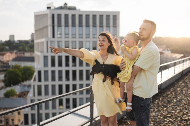 Duygusal esmer, ellerinde küçük bir daksund tutuyor ve bir adam bebeğe sarılırken işaret parmağıyla uzaklığı işaret ediyor. Sarı elbiseli sevgi dolu aile kızına temiz havada yeni şehirler gösteriyor..