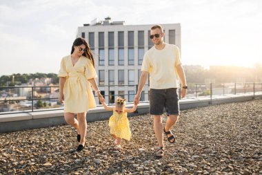 Yazlık giysiler içinde, çatıda taşlarla kaplı, dost canlısı beyaz bir aile. Gülümseyen eş ve koca dışarıda küçük bir çocuğun elini tutuyor. Ebeveynlik ve ilişki kavramı.