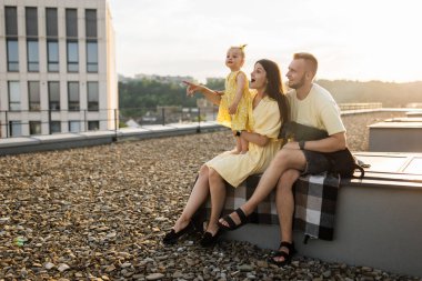 Dachshund köpeği açık alanda battaniyenin üzerinde oturan üç kişilik ailenin yan görüntüsü. Annesinin küçük kızına dışarıda bir şey göstermesine şaşırdım. Mutlu eş manzaranın ve gün batımının tadını çıkarıyor..
