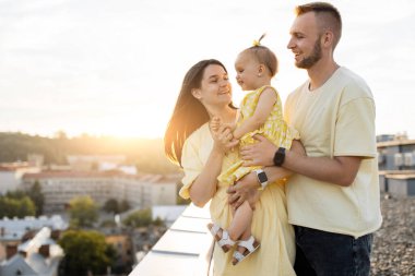 Örnek beyaz ebeveynler sevgili kızlarını kucaklıyor ve gökdelendeki terasta dinlenirken samimi bir şekilde gülümsüyorlar. Arka planda kentsel alanda şaşırtıcı yaz günbatımı.