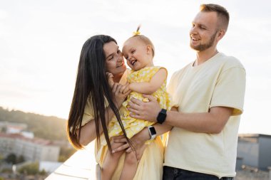 Örnek beyaz ebeveynler sevgili kızlarını kucaklıyor ve gökdelendeki terasta dinlenirken samimi bir şekilde gülümsüyorlar. Arka planda kentsel alanda şaşırtıcı yaz günbatımı.