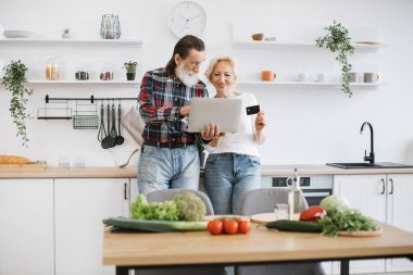 Yaşlı beyaz çift, modern geniş mutfak arka planında kahvaltı hazırlamak için dizüstü bilgisayar ve kredi kartı kullanıyor..