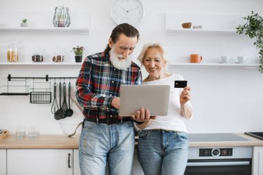 Mutlu yaşlı karı koca dizüstü bilgisayar ve kredi kartıyla modern ışık mutfağında lezzetli bir kahvaltı hazırlıyorlar..