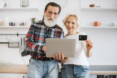 Dizüstü bilgisayara odaklanın. Ellerinde mutlu yaşlı bir koca ve eş var. Modern ışık mutfağında lezzetli bir kahvaltı hazırlamak için kredi kartı ödüyorlar..