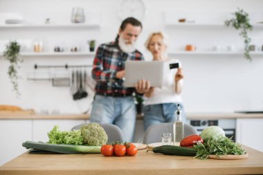 Masadaki sebzelere ve bitkilere odaklan. Yaşlı beyaz çift, modern geniş mutfak arka planında kahvaltı hazırlamak için dizüstü bilgisayar ve kredi kartı kullanıyor..
