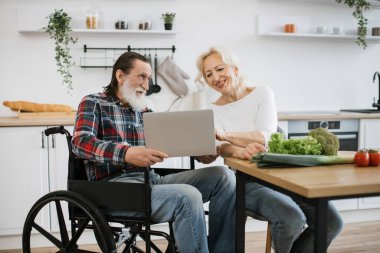 Gri sakallı yaşlı bir adam, tekerlekli sandalye kullanıcısı ve karısı elinde dizüstü bilgisayarla oturmuş internette lezzetli bir kahvaltı için ürünler seçiyorlar..