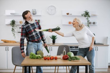 Mutlu kıdemli karı koca mutfakta eğleniyor, salatalık ve soğanı kılıç olarak kullanıp modern mutfakta sağlıklı salata hazırlıyor..