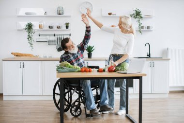 Tekerlekli sandalyedeki yaşlı adam ve karısı sağlıklı salata hazırlıyorlar, ince dilimler halinde sebze ve domatesleri bıçakla kesiyorlar ve birbirlerine beşlik çakıyorlar..