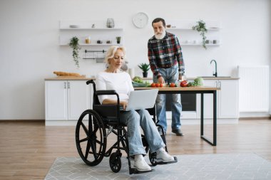 Yaşlı kadının tam boy portresi tekerlekli sandalyede oturur ve dizüstü bilgisayarda çalışırken gri sakallı kocası arka planda sağlıklı bir salata hazırlar..