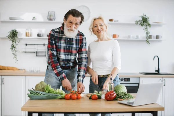 Caucasian old married man and wife prepare healthy salad, cutting thin slices of green vegetables and tomatoes with knife against background of spacious kitchen, looking at camera