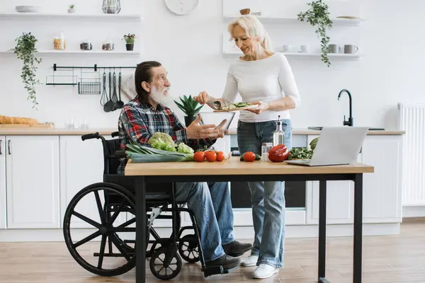 stock image Senior couple, man in wheelchair and elderly woman, making healthy salad. Male with gray beard holding bowl while his partner pours chopped vegetables.