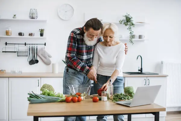 Positive retired couple cutting pepper while holding knife together in hands and putting pieces into glass bowl. Old family spends leisure time making healthy salad in modern kitchen.