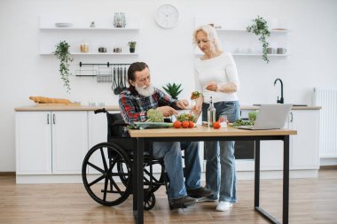 Engelli yaşlı çift modern aydınlık mutfakta kahvaltı hazırlamak için boş vakitlerini harcar. Yaşlı koca tekerlekli sandalyede ve karısı kasede sebze pişirirken lezzetli sağlıklı salata hazırlıyor..