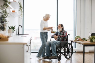 Mutlu yaşlı engelli çift panoramik pencereli modern mutfakta kahvaltı yapıyor. Sadık kadın, tekerlekli sandalyedeki adamı taze sağlıklı salatayla besler..