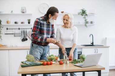 Gri sakallı yaşlı adam marul ve otları cam kaseye dökerken karısı da küçük parçalar halinde biber kesiyor. Yaşlı çift modern mutfakta taze sağlıklı salata hazırlıyor..