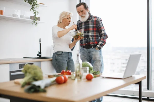 Happy old pensioner couple having breakfast in modern light kitchen with panoramic windows. Devoted elderly woman feeds man with gray beard with fresh healthy salad in glass bowl.