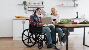 Yaşlı beyaz çift, büyüleyici bir eş ve tekerlekli sandalyede sakallı bir adam. Yemekleri kredi kartıyla ödüyor. Geniş mutfak arka planında dizüstü bilgisayar kullanıyor..