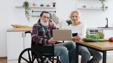 Yaşlı beyaz çift, büyüleyici bir eş ve tekerlekli sandalyede sakallı bir adam yemek parasını kredi kartıyla ödüyor. Bilgisayarla mutfağın arka planına bakıyor ve kameraya bakıyor.