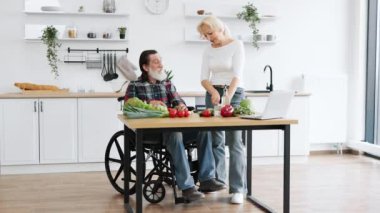 Tekerlekli sandalyede yaşlı ve evli bir adam sağlıklı salata hazırlar, sebze ve domatesleri ince ince dilimlere ayırır, geniş bir mutfak arka planında bıçakla keserler..