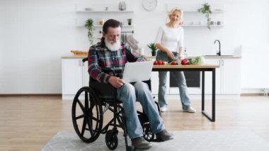 Gri sakallı yaşlı adam tekerlekli sandalyede oturup dizüstü bilgisayarda çalışırken karısı arka planda sağlıklı salata hazırlıyor..