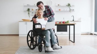 Tekerlekli sandalyedeki, laptoplu, yaşlı, çekici bir çift. Kocası, parlak modern mutfağın arka planında sarılıp kameraya bakıyor..