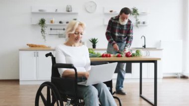 Yaşlı kadının yakın plan portresi tekerlekli sandalyede oturur ve dizüstü bilgisayarda çalışırken gri sakallı kocası arka planda sağlıklı bir salata hazırlar..