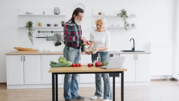 迷人的老夫妇夫妇一起在厨房里用新鲜蔬菜做健康的沙拉 白胡子男子将切碎的西红柿倒入伴侣手中的碗中 — 图库视频影像