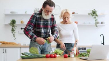 Günlük giysiler içinde yaşlı bir adam ve kadın marul ve otları cam kaseye döküyor. Yaşlı bir çift modern mutfakta taze sağlıklı salata hazırlıyor..