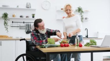 Özürlü yaşlı çiftin portresi modern mutfakta kahvaltı hazırlamakla zaman harcıyor. Yaşlı koca tekerlekli sandalyede ve karısı kasede sebze pişirirken lezzetli sağlıklı salata hazırlıyor..