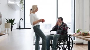 Mutlu emekliler, evli, gri sakallı ve tekerlekli sandalyeli. Parlak, modern bir mutfakta lezzetli bir kahvaltıdan sonra bardaklarda sıcak içecekler tokuşturuyorlar..