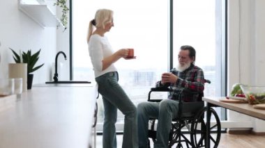 Mutlu emekliler, evli, gri sakallı ve tekerlekli sandalyeli. Parlak, modern bir mutfakta lezzetli bir kahvaltıdan sonra bardaklarda sıcak içecekler tokuşturuyorlar..