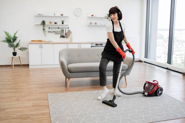 Full length portrait of cleaning service employee listens to music and dances while cleaning bright and spacious apartment. Positive young cleaners vacuuming rug of stylish modern studio kitchen.
