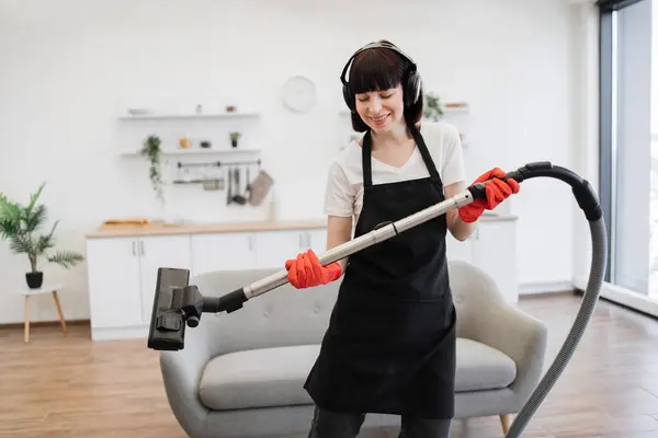 Cleaning service employee listens to music in headphones and dances while cleaning bright and spacious apartment. Positive young cleaners vacuuming rug of stylish modern studio kitchen.