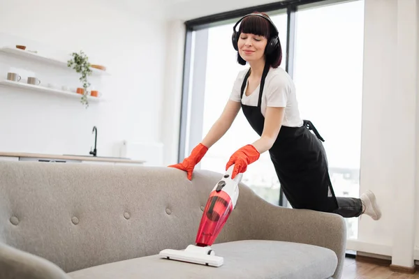 Portrait of happy smiling employee of cleaning company cleaning in bright, spacious home kitchen. Professional young cleaner vacuuming sofa with portable cordless vacuum cleaner.