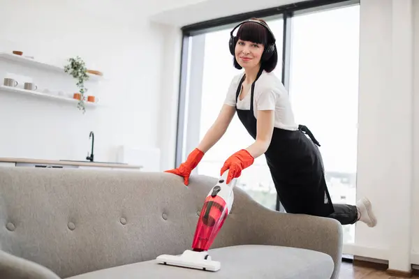 Portrait of happy smiling employee of cleaning company cleaning in bright, spacious home kitchen posing to camera. Professional young cleaner vacuuming sofa with portable cordless vacuum cleaner.